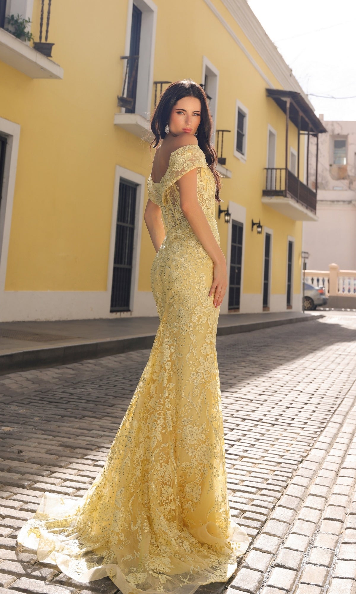 A woman wearing the Off-the-Shoulder Long Lace Prom Dress C1455 stands on a cobblestone street lined with yellow buildings and balconies, glancing over her shoulder at the camera.